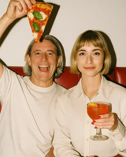 Man and woman posing with a slice of pizza and an aperol